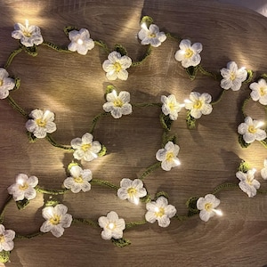 Puede incluir: Una guirnalda de flores de ganchillo con pequeñas luces LED. Las flores blancas tienen centros amarillos y están conectadas por una vid verde. Las luces están encendidas, creando un brillo cálido. La guirnalda está colocada sobre una superficie de madera.