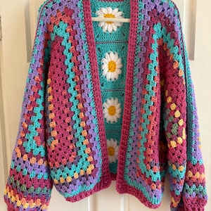 May include: A colourful, open-front crochet cardigan in a granny square design. The colour scheme includes pinks, purples, teal, and yellow. The central panel features daisy appliqués. The cardigan is displayed on a white hanger against a white door.