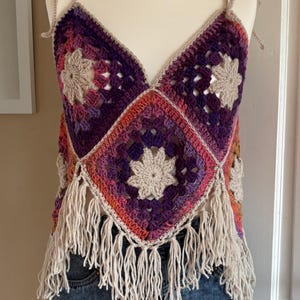 May include: A crocheted top with a geometric pattern in shades of purple, pink, and orange. The top has a V-neckline and is trimmed with fringe.