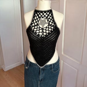 May include: Black crochet halter top with a gray star applique. The top has a halter neck and a triangular shape. The edges are trimmed with gray yarn. The top is displayed on a mannequin.