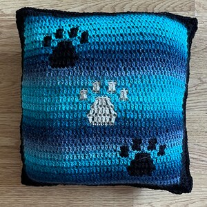 May include: A blue and black crocheted pillow with three paw prints. The paw prints are in a white, black, and gray color scheme.