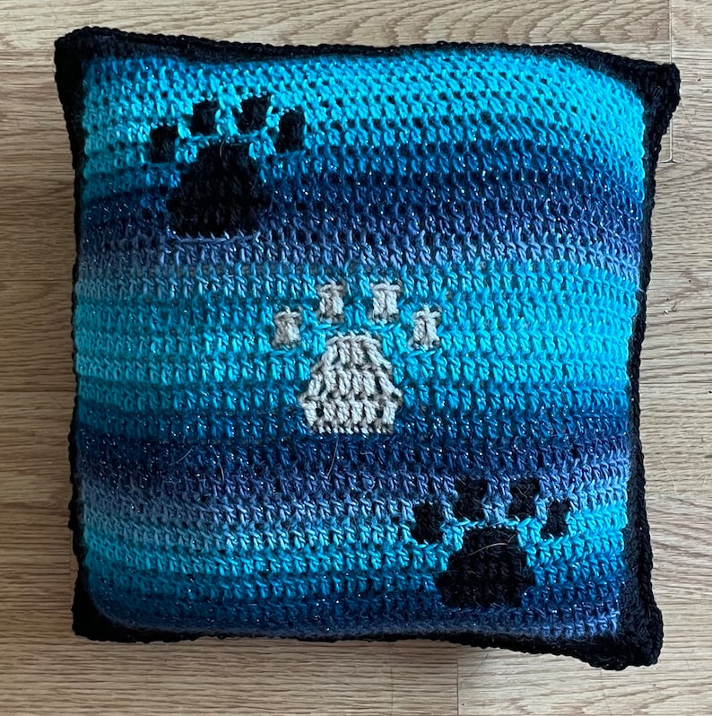 May include: A blue and black crocheted pillow with three paw prints. The paw prints are in a white, black, and gray color scheme.