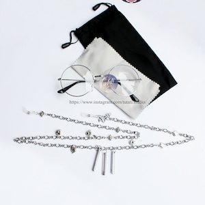 May include: A set of silver-toned glasses chains with decorative charms. The chains are laid out on a white surface with a pair of round, clear-framed glasses and a black glasses case. The text "UKI VIOLETA" is visible at the top of the image. The text "https://www.instagram.com/tutati.studio/" is visible below the glasses.