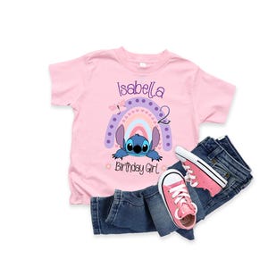 May include: Pink t-shirt with a blue Stitch character from Lilo & Stitch peeking out from behind a rainbow with pink, purple, and blue stripes. The shirt says "Isabella 2 Birthday Girl" in purple text.