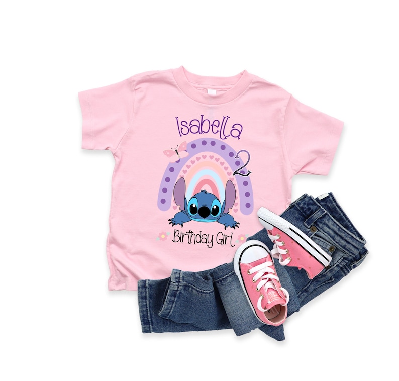 May include: Pink t-shirt with a blue Stitch character from Lilo & Stitch peeking out from behind a rainbow with pink, purple, and blue stripes. The shirt says "Isabella 2 Birthday Girl" in purple text.