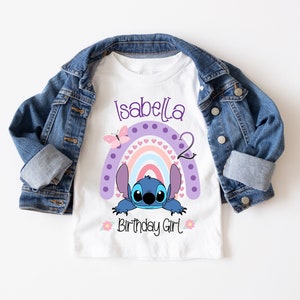 May include: White t-shirt with a purple, pink, and blue rainbow with a blue Stitch character peeking out from behind it. The text "Isabella 2 Birthday Girl" is printed on the shirt.