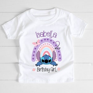 May include: White t-shirt with a purple, pink, and blue rainbow with a blue Stitch character peeking out from behind it. The text "Isabella 2 Birthday Girl" is printed on the shirt.
