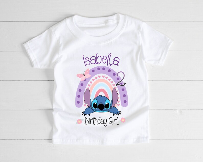 May include: White t-shirt with a purple, pink, and blue rainbow with a blue Stitch character peeking out from behind it. The text "Isabella 2 Birthday Girl" is printed on the shirt.