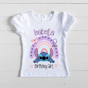 May include: White t-shirt with a purple, pink, and blue rainbow with white hearts, a blue Stitch character peeking out from behind the rainbow, and the text "Isabella 2 Birthday Girl".