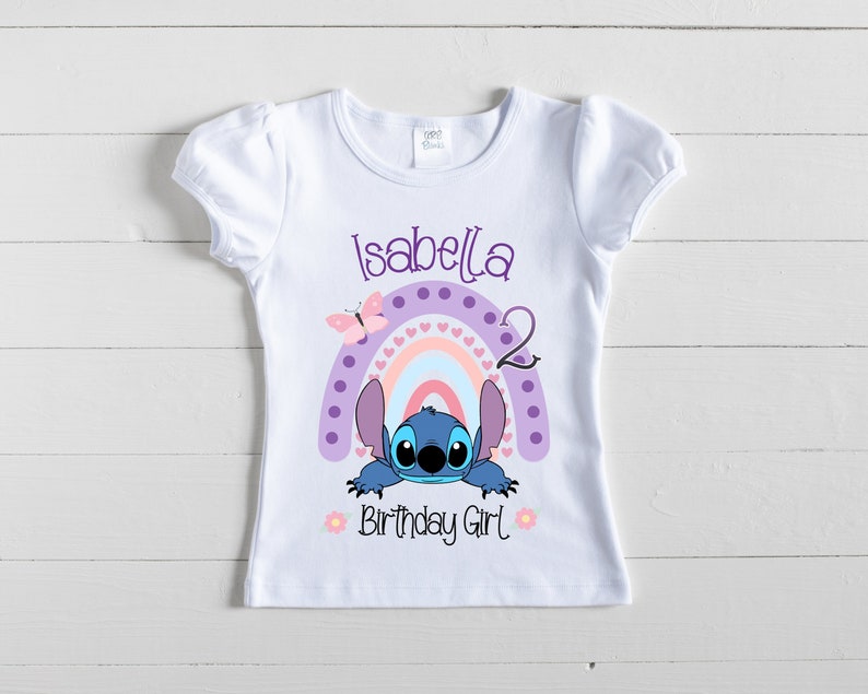 May include: White t-shirt with a purple, pink, and blue rainbow with white hearts, a blue Stitch character peeking out from behind the rainbow, and the text "Isabella 2 Birthday Girl".