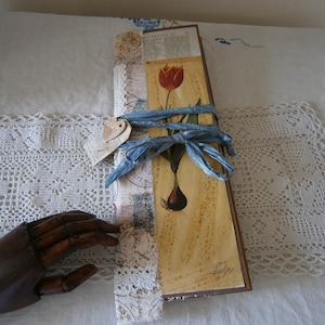 May include: A long, rectangular journal tied with a light blue ribbon. The journal has a tulip illustration and a small heart-shaped tag. The background includes lace and vintage-style paper.