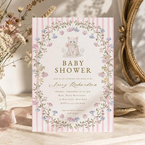 May include: A rectangular baby shower invitation card featuring a light pink and white vertical striped background. The centre displays a kitten illustration surrounded by a floral wreath. Text reads: BABY SHOWER, Avery Richardson, Sunday, September 26 at 2pm, Rose Heart Restaurant, 32 Rose Lane, New York, RSVP to 555-768-8768, registered at amazon and target.