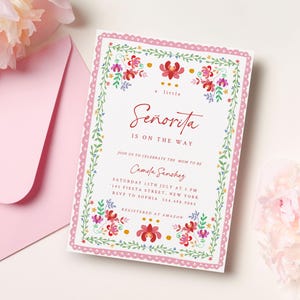 Könnte beinhalten: Eine weiße Einladungskarte mit einem rosa, gewellten Rand und floralen Akzenten. Der Text lautet "a little Señorita is on the way". Enthält Eventdetails und RSVP-Informationen. Ein rosa Umschlag und Blumen sind im Hintergrund.