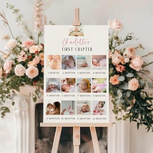 May include: A wooden easel holds a sign with the text "Charlotte's FIRST CHAPTER." The sign showcases a collage of baby photos, each labelled with a month from 1 to 12. The sign is decorated with floral elements.