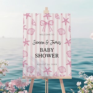 以下が含まれることがあります： 木製のイーゼルに立てられたベビーシャワーのウェルカムサイン。ピンクと白のストライプの背景に、貝殻とヒトデのイラストが描かれています。テキストは「Welcome to Sienna & James BABY SHOWER August 26, 20XX」と書かれています。