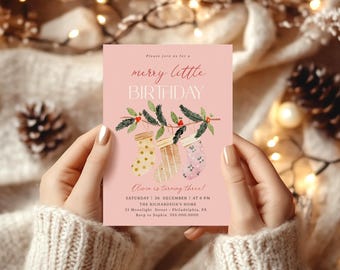 May include: A rectangular birthday invitation card on a light pink background featuring the text "merry little BIRTHDAY" and "Olivia is turning three!". The design includes watercolor illustrations of three hanging stockings.