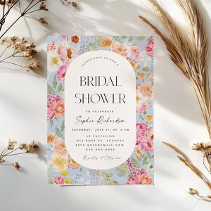 Pu&ograve; includere: Un invito a un bridal shower floreale con sfondo blu chiaro e cornice ovale bianca. Il testo recita "Si prega di unirsi a noi per un bridal shower per celebrare Sophia Richardson, sabato 17 luglio, alle 18:00, Le Pavillion, 315 Rose St, Augusta, RSVP rose@gmail.com, 555.000.0000, Registro su Etsy."