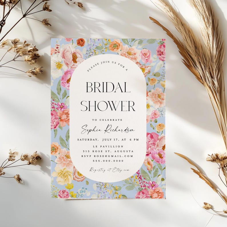 Pu&ograve; includere: Un invito a un bridal shower floreale con sfondo blu chiaro e cornice ovale bianca. Il testo recita "Si prega di unirsi a noi per un bridal shower per celebrare Sophia Richardson, sabato 17 luglio, alle 18:00, Le Pavillion, 315 Rose St, Augusta, RSVP rose@gmail.com, 555.000.0000, Registro su Etsy."