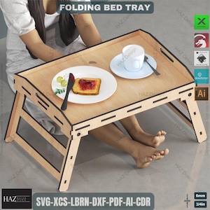 May include: A wooden folding bed tray with a light wood finish. The tray has a black border and is supported by two foldable legs. The tray is shown with a plate, a cup, a knife, and a piece of toast with jam on it. The text "SVG-XCS-LBRN-DXF-PDF-AI-CDR" is printed on the tray. The tray is 6mm (1/4in) thick.