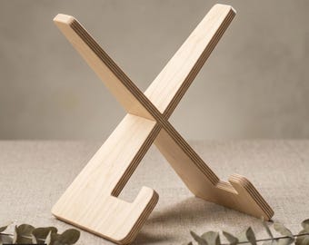 May include: A light-colored wooden stand with an X-shaped design. The stand has a base with a curved lip to hold an item. The text "SVG-DXF-PDF-AI-CDR" is visible. The stand is made of layered wood.