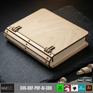 Può includere: Una scatola rettangolare in legno chiaro con coperchio incernierato e venature visibili. La scatola presenta una costruzione a strati con accenti scuri sulle cerniere e sui bordi. Un'etichetta indica uno spessore di 3mm.