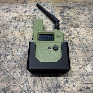 May include: An olive green electronic device with a black antenna and a small screen displaying text and graphics. The device is in a black charging cradle. The screen reads "been yesterday" and "Hope Aware 1".