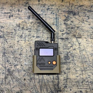 May include: A black electronic device with a screen, antenna, and orange buttons, held in a tan-colored bracket. The device has the text "PWRD" printed on it. The background is a textured, wooden surface.