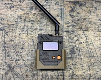 May include: A black electronic device with a screen, antenna, and orange buttons, held in a tan-colored bracket. The device has the text "PWRD" printed on it. The background is a textured, wooden surface.