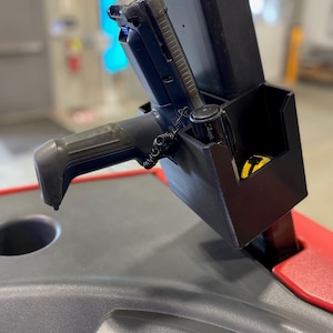 May include: A black plastic holder with a black and gray handheld scanner and a black marker. The holder is mounted on a gray surface.