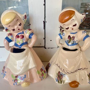 May include: Two ceramic figurines of Dutch girls in traditional attire. The figures are painted in pastel colors with floral accents. Each figurine has a skirt, apron, and a hat. The figurines are approximately 6 inches tall.