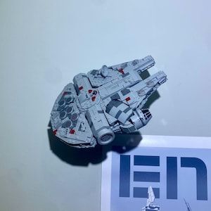 May include: A light gray model of the Millennium Falcon spaceship, featuring intricate details and red accents. The model is constructed from interlocking blocks, showcasing the iconic design of the spacecraft. The model is a collectible item.