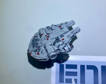 May include: A light gray model of the Millennium Falcon spaceship, featuring intricate details and red accents. The model is constructed from interlocking blocks, showcasing the iconic design of the spacecraft. The model is a collectible item.