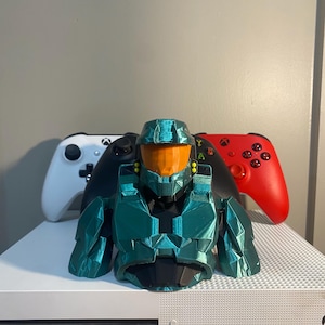 May include: A teal green 3D printed bust of a Master Chief Halo character, with an orange visor, standing on a white Xbox console with two controllers, one white and one red.
