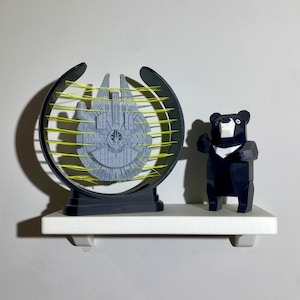 Könnte beinhalten: Ein graues Millennium Falcon-Modell ist in einem schwarzen und gelben kreisförmigen Rahmen eingeschlossen, der auf einem weißen Regal ausgestellt ist. Daneben steht eine schwarz-weiße geometrische Bärenfigur.