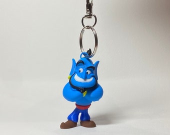 May include: A blue Genie keychain with a silver clasp and key ring. The Genie figure has a smiling expression, crossed arms, and is wearing blue pants, brown shoes, a red belt, and a black necklace. The keychain is suspended against a white background.