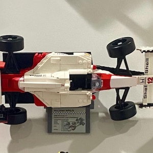 May include: A red, white, and black Lego model of a McLaren Formula 1 race car. The model has black wheels and the Shell logo. A small gray box with text is beneath the car.
