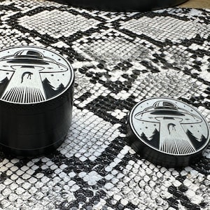 May include: Two black metal grinder with a silver UFO design. The UFO is flying over a forest with stars in the background.