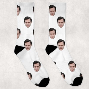 May include: A pair of white socks featuring a repeating pattern of a person's face. The socks have black heels and toes. The faces are evenly spaced across the white fabric.