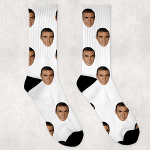 May include: A pair of white socks with a black heel and toe. The socks feature a repeated pattern of a man's face.
