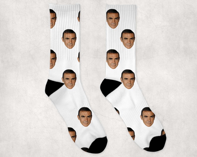 May include: A pair of white socks with a black heel and toe. The socks feature a repeated pattern of a man's face.