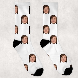 Op de afbeelding: Een paar witte sokken met een herhalend patroon van een gezicht. De sokken hebben zwarte hakken en tenen.