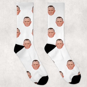 Peut inclure: Une paire de chaussettes blanches avec un motif répété d'un visage d'homme. Les chaussettes sont noires au niveau des orteils et du talon.