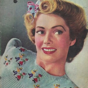 May include: A woman with blonde hair styled in pin curls wears a light blue sweater with a colorful embroidered design of people dancing. She is smiling and wearing a blue and white floral hair bow.