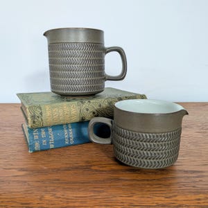 May include: Two ceramic pitchers with a dark gray finish and a textured herringbone pattern on the lower half. Each pitcher features a smooth upper rim, a small pouring spout, and a side handle. The interior of the pitchers is white.