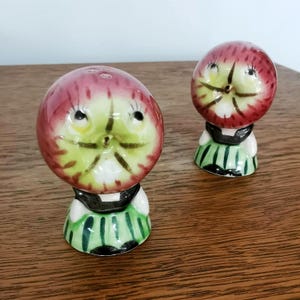 May include: Two ceramic apple-shaped salt and pepper shakers. The apple tops are red with green and yellow faces. They have black collars and green and white striped skirts. The shakers are on a wooden surface.