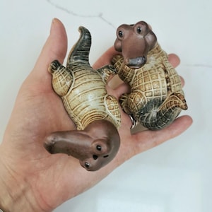 May include: Two ceramic alligator figurines. One is upside down, the other upright. The alligators are tan and brown with textured details. The figurines are held in a hand.