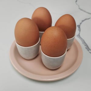 May include: Four brown eggs in white ceramic egg cups, presented on a light pink plate. The eggs are a warm brown colour, and the cups have a textured, matte finish. The plate is round with a slightly raised edge.
