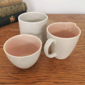 May include: Three ceramic pieces: a small bowl, a cylindrical cup, and a mug with a handle. The exteriors are a textured off-white, while the interiors of the bowl and mug are a soft pink. The items are arranged on a wooden surface.