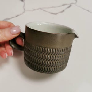 May include: A brown ceramic jug with a white interior and a textured chevron pattern. The jug has a handle and a spout.
