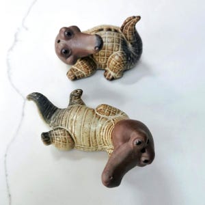 May include: Two ceramic alligator figurines featuring a textured, cream-colored body with a grid-like pattern and a smooth, dark brown head. Each alligator has small black eyes and a curved tail, with one positioned upright and the other lying flat.
